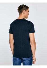 Mladić okrenut leđima nosi jednostavnu tamnoplavu majicu i plave traperice, na svijetlosivoj pozadini.