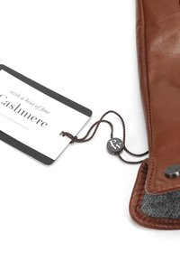 Hnědý kožený výrobek se šedým interiérem, který má štítek s nápisem "Cashmere" velkým písmem a kulatým logem, které je uvázané hnědým provázkem.