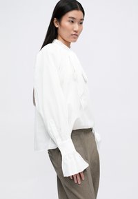 Camicia bianca a maniche lunghe con colletto alto e polsini arricciati, caratterizzata da un delicato dettaglio a fiocco al colletto. Indossata con pantaloni marroni a fantasia.