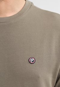 Sweatshirt verde oliva con scollo rotondo e polsini a costine. Presenta un patch circolare con logo in blu, rosso e nero sul petto.