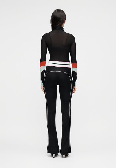 Γυναίκα που στέκεται με την πλάτη, φορώντας μαύρη διάφανη μπλούζα μακρυμάνικη με έγχρωμες μανσέτες και εφαρμοστά μαύρα παντελόνια με λευκή ρίγα και ψηλοτάκουνα.