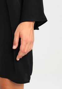 Svart plagg med ett flödande mönster och breda ärmar. En hand är placerad nära fållen och visar det mjuka, lätta tygets textur.