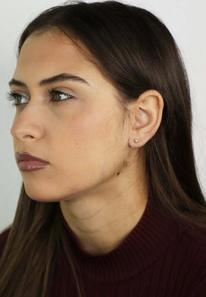 Młoda kobieta o długich brązowych włosach, ubrana w ciemnoczerwony golf, z małym kolczykiem w sztyfcie, spoglądająca na bok na jednolitym białym tle.