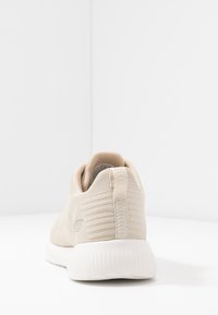 Sneaker lavorato a maglia beige con suola bianca a coste vista da dietro, con una linguetta e un piccolo logo sul lato.