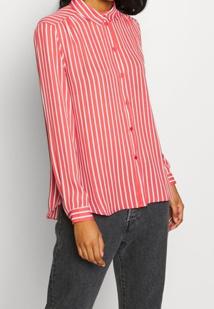 Vrouw draagt een rood hemd met lange mouwen met witte verticale strepen en zwarte jeans, staand tegen een effen lichte achtergrond.