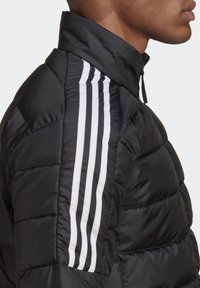 Homme portant une veste matelassée noire avec un col montant et trois rayures blanches courant le long de l'épaule et du bras.