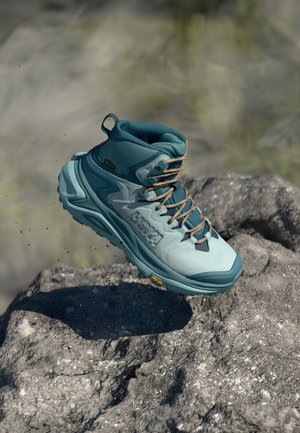 Sapatilha de caminhada azul clara com um cano alto, parte superior texturizada, atacadores castanhos e sola acolchoada. Marca Vibram visível na sola.