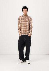 Brun og kremfarget rutete skjorte med knapper og en lomme, kombinert med løse svarte jeans og hvite joggesko. Modellen har på seg en svart lue.