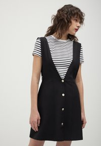 Robe noire en coupe A avec un décolleté plongeant et des boutons décoratifs, portée sur un haut à rayures noir et blanc. Tissu lisse, design décontracté.