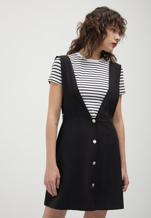 Schwarzes A-Linien-Kleid mit tiefem Ausschnitt und Knopfdetails, über einem schwarz-weißen gestreiften Oberteil getragen. Glatter Stoff, lässiges Design.