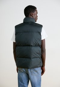 Sort polstret vest med høj krave, prydet med quiltet design og letvægtsmateriale, båret over en hvid skjorte og kombineret med blå jeans.