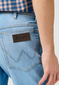 Jeans Wrangler blu chiaro con una toppa in pelle marrone, caratterizzati da cuciture gialle e un distintivo motivo a zigzag sulla tasca posteriore.