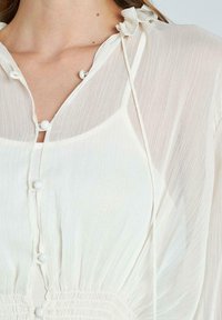 Lange witte blouse van lichtgewicht, doorzichtig materiaal met verticale plooien, voorzien van een knoopsluiting aan de voorkant en zachte nekstrikjes.