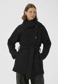 Černý kabát z vlněné směsi s širokým límcem, dvouřadým knoflíkováním, páskem a oversized rukávy. Má strukturovaný povrch.