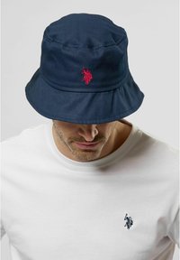 Marinblå bomullsbucket-hatt med en röd broderad logga frampå, med en bred skärm och en strukturerad krona. Bärs med en vit t-shirt.