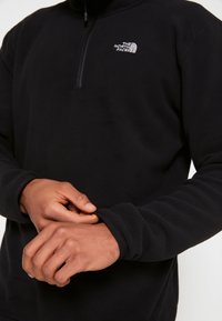 Pullover in fleece nero con zip a un quarto, caratterizzato da un logo sulla parte superiore del petto sinistro e polsini arrotolati per una vestibilità aderente. Tessuto morbido.