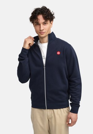 Sudadera con cremallera en color navy, hecha de un tejido suave, que cuenta con un cuello alto y un parche con un logo rojo. Se muestra usada sobre una camiseta blanca.