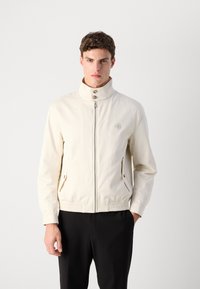 Light beige jacket with a stand collar, front zipper, and two side pockets. Features a gathered waistband and a button accent on the chest.