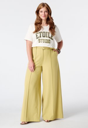 Vrouw met lang bruin haar die een wit "Etoile Studio" overhemd en hooggetailleerde, wijde gele broek draagt, staand met één hand in de zak.