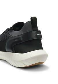 Baskets de sport noires avec une tige en mesh texturé, des accents en cuir, un onglet au talon et une semelle extérieure en caoutchouc blanc avec un motif de bande de roulement.