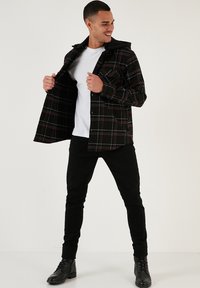 Camicia scozzese nera e rossa con cappuccio, indossata aperta su una t-shirt bianca, abbinata a pantaloni neri e stivali neri stringati.