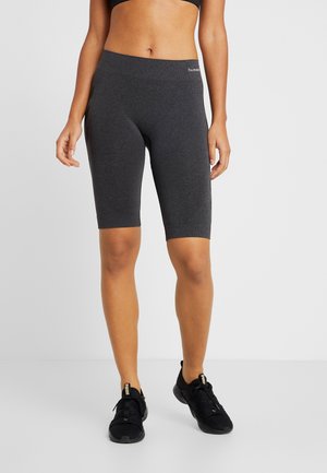 Kvinde iført mørkegrå tætsiddende shorts til knæet og sorte sportssko, stående mod en ensfarvet lys baggrund.