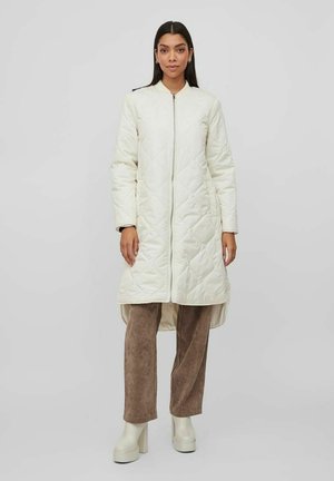 Gepolsterte, knielange, cremefarbene Outerwear mit Reißverschluss vorne, langen Ärmeln und seitlichen Schlitzen. Das Model trägt braune Hosen und klobige, hellfarbene Stiefel.