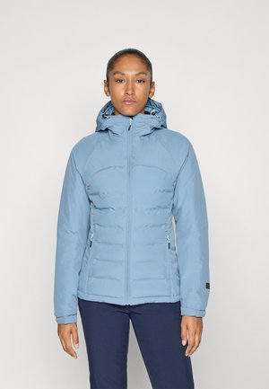 Femme portant une veste à capuche matelassée bleu clair avec des poches zippées, se tenant devant un fond uni clair.