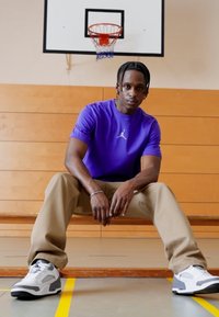 Camiseta de manga corta morada con un logo, pantalones beiges y zapatillas grises con acentos negros, sentado en un banco de madera en un gimnasio. Aro de baloncesto al fondo.