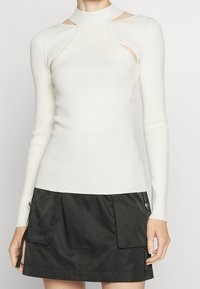 Maglione bianco a coste con collo alto, maniche lunghe e spacchi laterali. Abbinato a una gonna nera al ginocchio con tasche cargo.