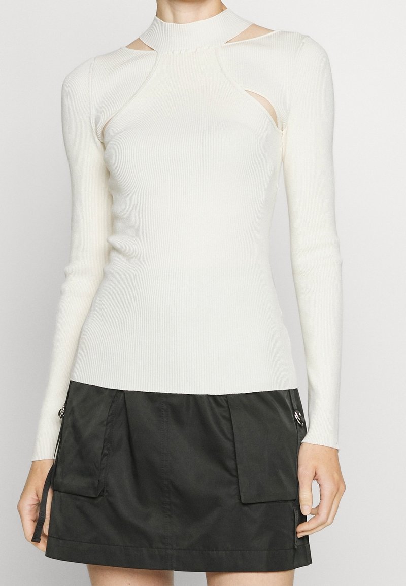 Maglione bianco a coste con collo alto, maniche lunghe e spacchi laterali. Abbinato a una gonna nera al ginocchio con tasche cargo.