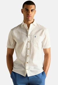 Īsām piedurknēm, balta lina krekls ar pogām priekšpusē, brūnas pogas un zils logotips uz krūšu kabatas. Gluda tekstūra un brīva piegriezuma.