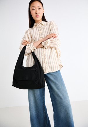 Kobieta w beżowej, w paski, zapinanej na guziki koszuli i szerokich niebieskich dżinsach, trzymająca dużą czarną torbę na ramię na białym tle.
