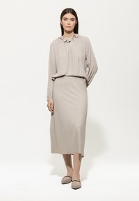 Frau, die eine langärmlige, beigefarbene Bluse mit Rüschen, einen Midikleid-Rock und beigefarbene flache Schuhe trägt, vor einem weißen Hintergrund steht.