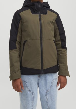 Hombre con chaqueta con capucha de color verde oliva y negro con cremallera frontal y vaqueros azul claro con costuras detalladas.