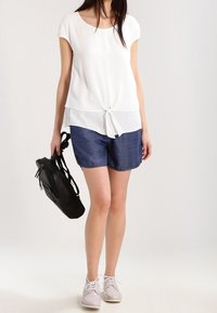 Valkoinen lyhythihainen paita, jossa on solmittu etuosa, yhdistettynä tummansiniset pilkkushortsit. Mallilla on musta laukku kädessä ja vaaleat kengät jalassa.