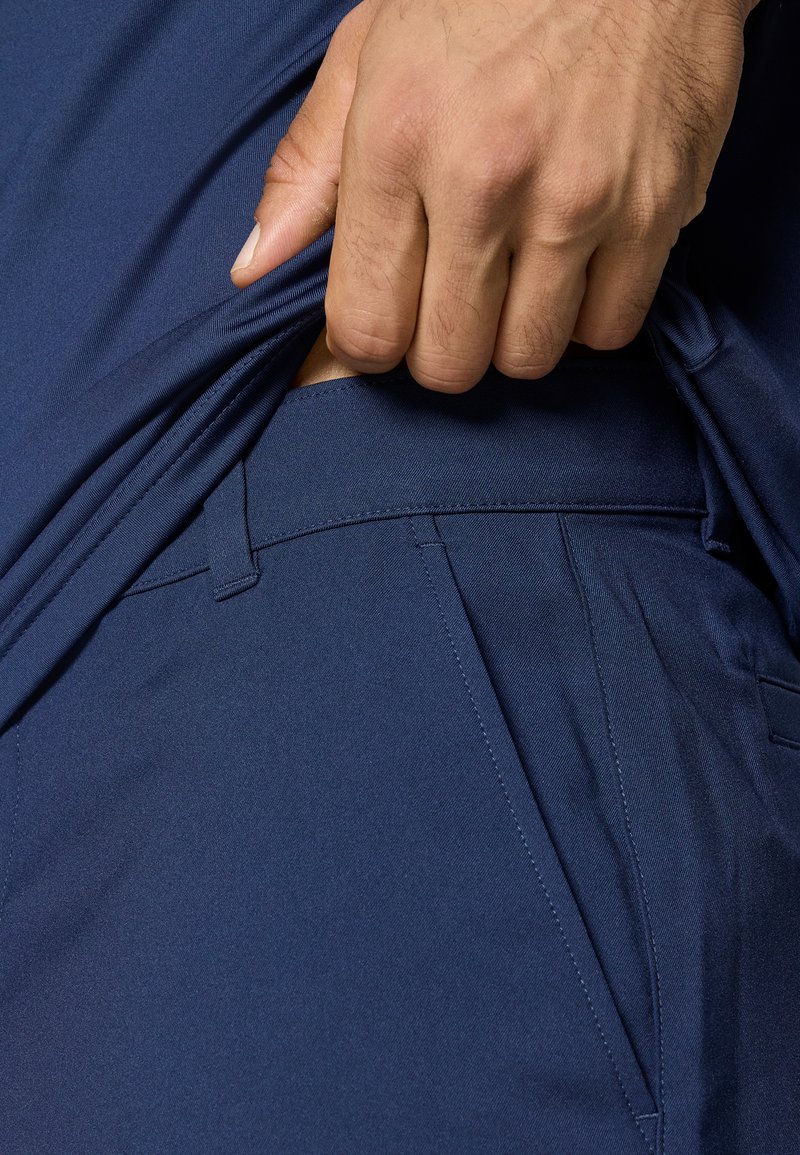 Marineblaue Sporthosen mit einer glatten Textur, ausgestattet mit einem flachen Frontbund und Seitentaschen, teilweise gezeigt mit einer Hand, die den Bund greift.