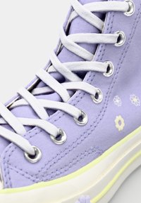 Purple canvas high-top sneaker with white laces, metallic eyelets, and yellow accents; features small floral designs on the side.