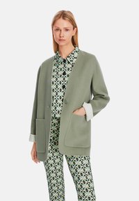 Hellgrüner, weicher Wollmischung langer Cardigan mit einem einzelnen Knopf, vorderen Taschen und umgeschlagenen Manschetten, getragen über einer gemusterten Bluse und Hose.