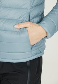 Lyseblå puffjakke med horisontalt kviltet mønster, der har en sidelomme og en tætsiddende pasform ved taljen, parret med sorte bukser.