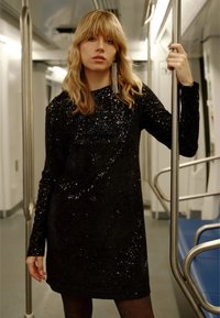 Czarna sukienka z cekinami z długimi rękawami, wyposażona w fakturowany wzór. Modelka trzyma rurkę w metro, wewnątrz pociągu, ukazując odbicia.