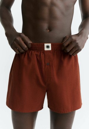 Donkere persoon die de tailleband vasthoudt van losse rode geruite boxershorts met een zwarte knoop en een wit label.