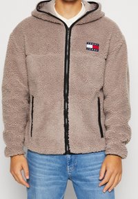 Giacca con cappuccio beige pelosa, con zip frontale, dettagli neri e patch "Tommy Jeans" sul petto. Due tasche laterali con zip.