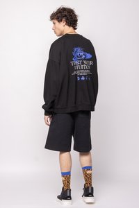 Sudadera negra oversized con estampado y texto azul en la parte posterior, combinada con pantalones cortos negros y calcetines de leopardo naranja. Se llevan zapatillas negras.
