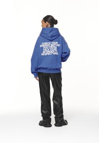Blue oversized hoodie in cotton blend, featuring large white text on the back. Paired with black faux leather pants and black shoes.