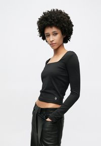 Haut noir à manches longues avec un col carré, en tissu lisse. Présente une coupe ajustée et un petit logo sur l'ourlet. Associé à un pantalon en cuir.