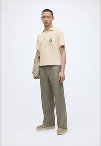 Homme debout portant un polo beige à manches courtes avec broderie d'arbre, pantalon gris à jambes larges, chaussures marron sans lacets, tenant une veste beige.