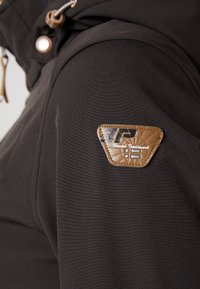 Chaqueta de tela negra con un parche de logo texturizado en marrón en el hombro; cuenta con elementos metálicos y cordón ajustable en la capucha.
