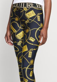 Svarta leggings med ett glänsande mönster av guld kedja och emblem. Den elastiska midjebandet har "COUTURE" i metallisk text. Slät textur.