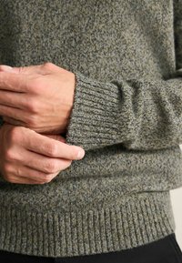Mörkgrön stickad tröja med ett texturerat mönster. Ribbad mudd och fåll. Händer håller tröjans ärm och visar tygets tjocklek.